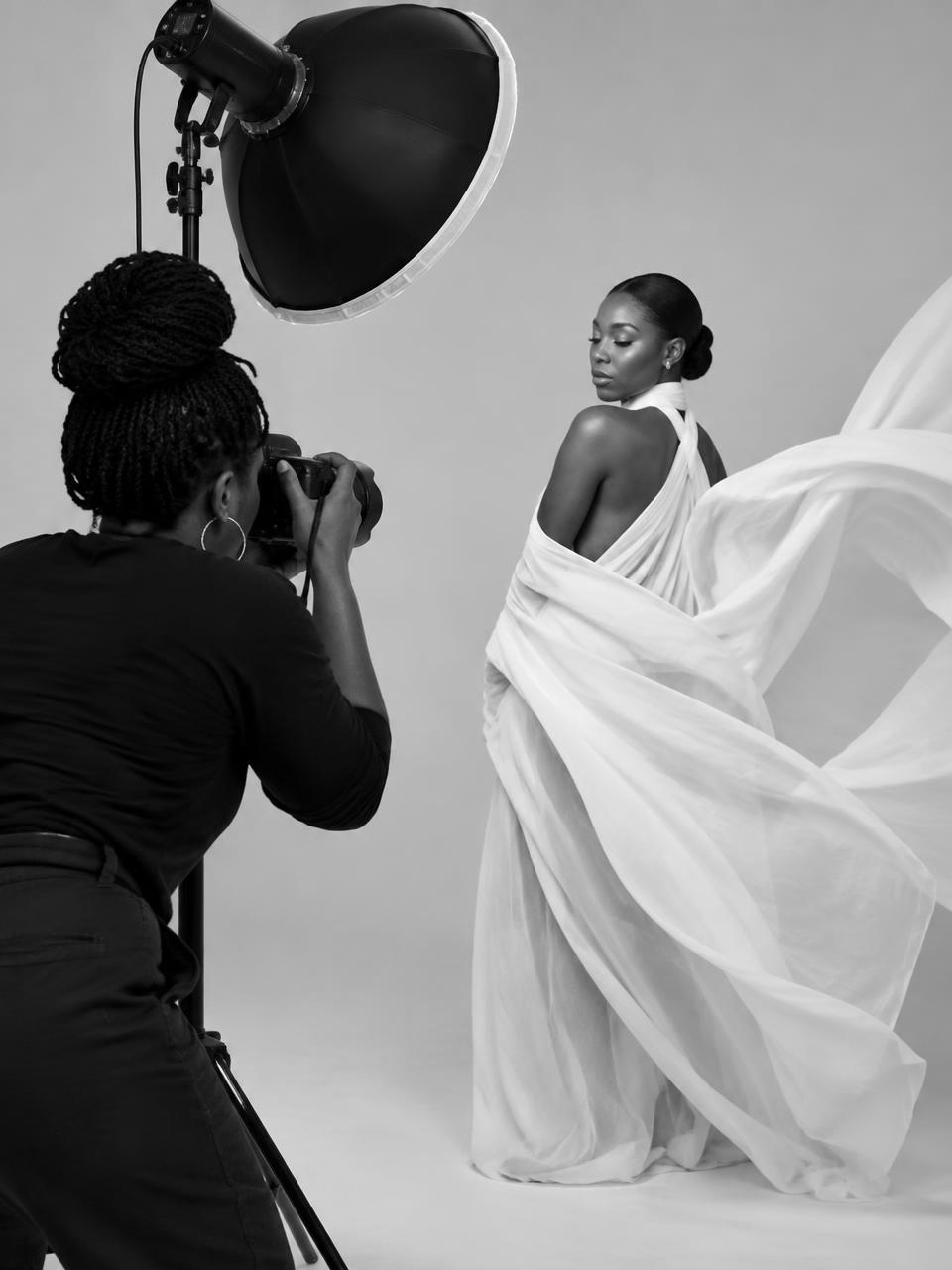 Black and white studio scene of a photographer behind a strobe with a model in flowing white fabric